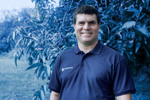 A man wearing a navy blue AGRAYA polo shirt stands outdoors in front of leafy plants, smiling at the camera.