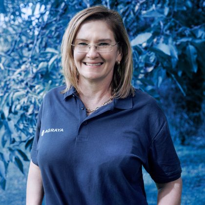 Elmé Coetzer-Boersma wearing glasses and a navy polo shirt with "AGRAYA" embroidered stands outdoors near green foliage, smiling at the camera.