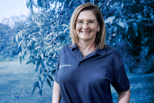 A woman wearing glasses and a navy polo shirt with "AGRAYA" embroidered stands outdoors near green foliage, smiling at the camera.