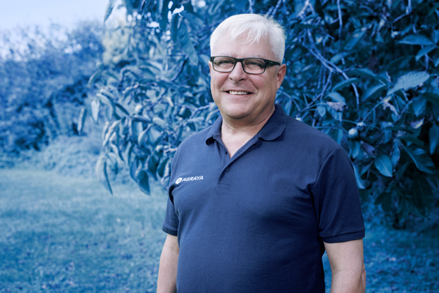 A man with short white hair and glasses stands outdoors, smiling, wearing a dark blue polo shirt with a logo on the chest. Green trees and grass are in the background.