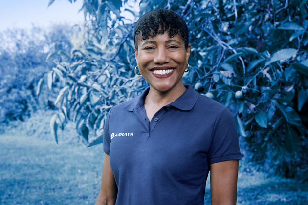 A person wearing a navy blue polo shirt with the Agraya logo smiles while standing outdoors near leafy trees.