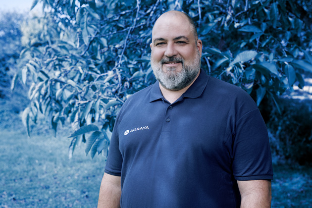 A man with a beard wearing a navy blue polo shirt with the logo "AGRAYA" stands outdoors in front of leafy trees.
