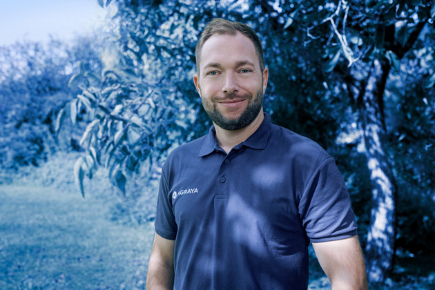 A man with a beard wearing a navy polo shirt stands outdoors in front of trees and greenery, smiling at the camera.