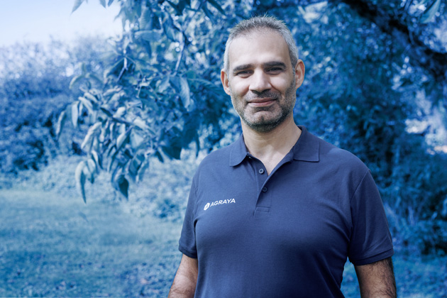 A man wearing a navy blue polo shirt with "AGRAYA" stands outdoors in front of leafy trees, smiling at the camera.