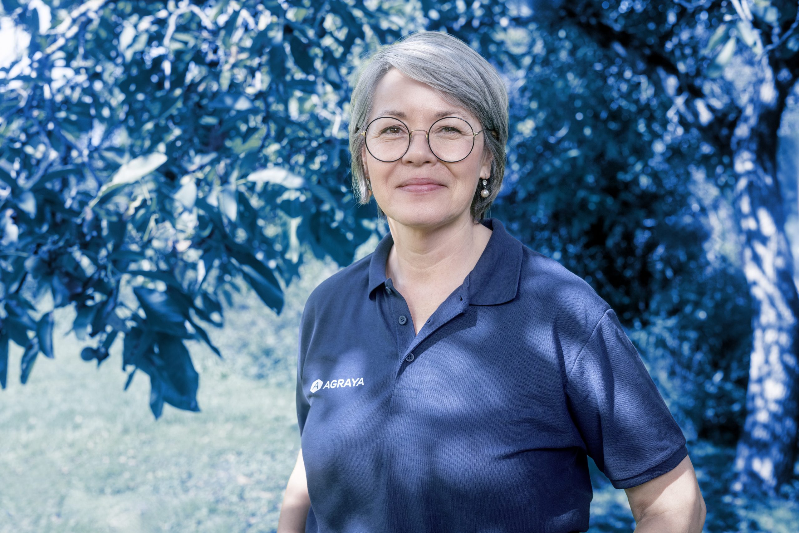 A woman wearing a navy blue polo shirt with the Agraya logo smiles while standing outdoors near leafy trees.