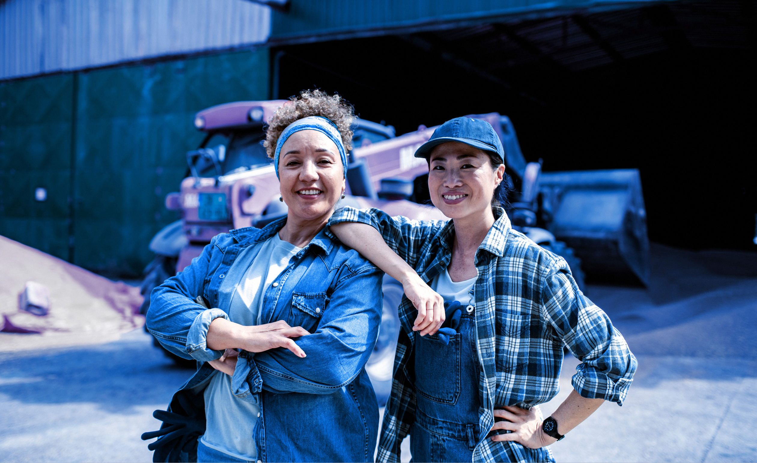 Two women in denim clothing stand smiling in front of a tractor and a barn